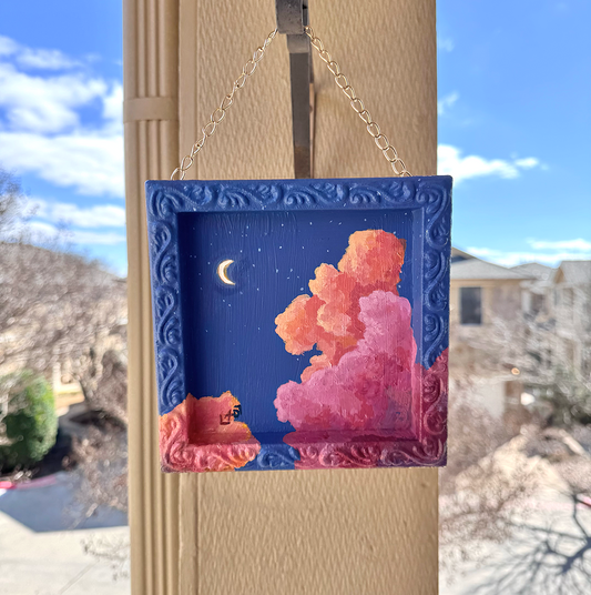Decorative framed artwork with pink and orange clouds hanging on an outdoor column.
